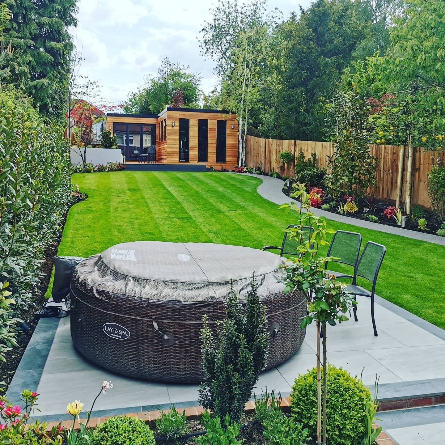 A striped lawn leading to a cedar-clad garden room, with a covered hot tub and metal patio chairs on the stone patio in the foreground