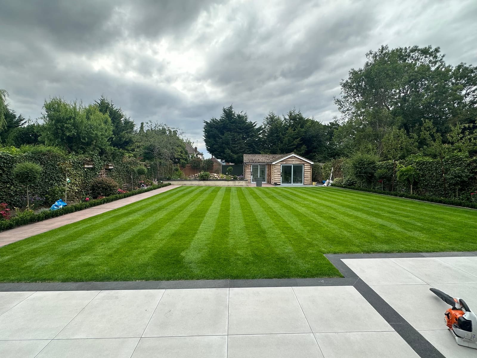 Striped lawn bordered by hedging leading to a garden room, with a paved patio in the foreground
