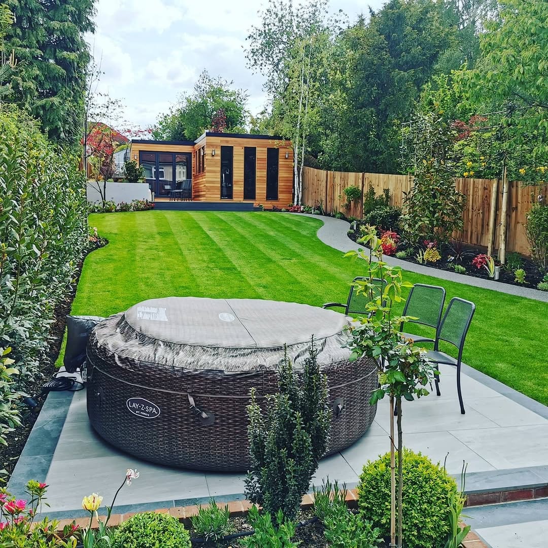 A striped lawn leading to a cedar-clad garden room, with a covered hot tub on the patio in the foreground