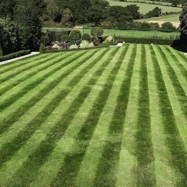 A striped lawn leading to a hot tub gazebo and topiary garden, bordered by a green estate fence with rolling countryside beyond