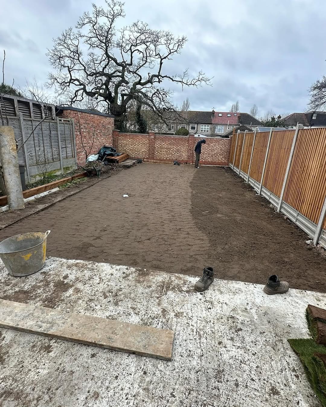 A garden with bare, raked soil ready for turf, a worker checking the ground, boots and tools left on the concrete path
