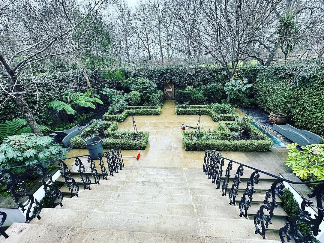 An elegant formal garden with clipped box parterres and tree ferns, reached by stone steps, with a swimming pool to one side